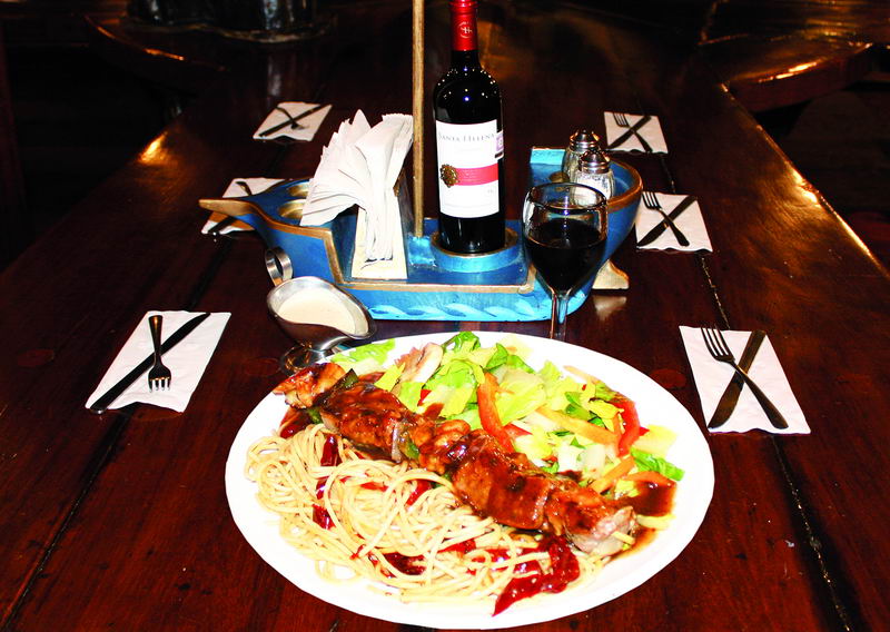 Dinner plate served aboard the ship: grilled meat skewers over spaghetti with barbecue sauce and a side salad, with a bottle of red wine and a filled glass in a boat-shaped napkin holder on the dark wood table. Dim dining-show lighting.