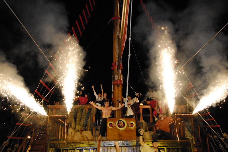 Nighttime pirate show finale on the ship's deck with bright pyrotechnic sparkler fountains shooting up on both sides of the mast while shirtless and costumed performers raise their arms in celebration. Dramatic stage lighting against a black sky.