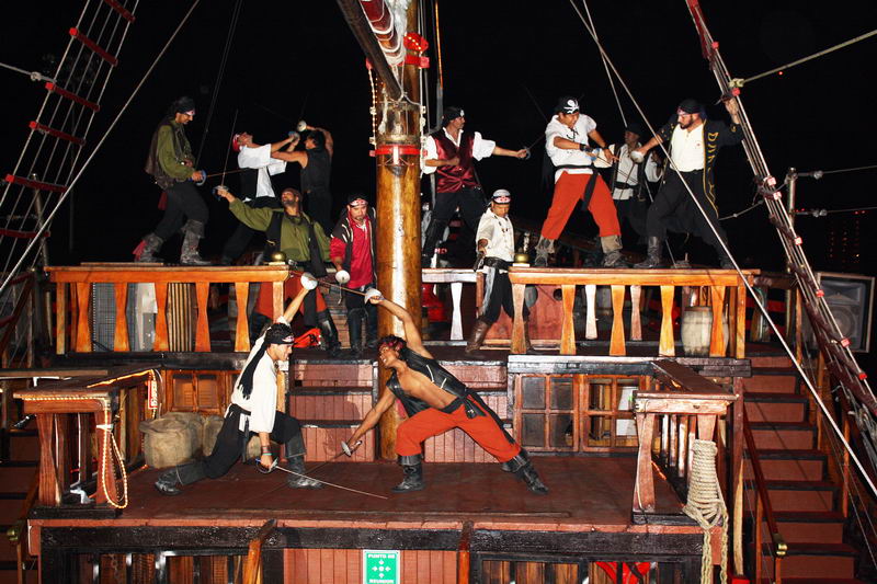 Nighttime pirate show scene with numerous costumed performers staging a sword fight across the upper and lower decks of the galleon under stage lighting. Two swordsmen duel in the foreground; the black night sky is behind.