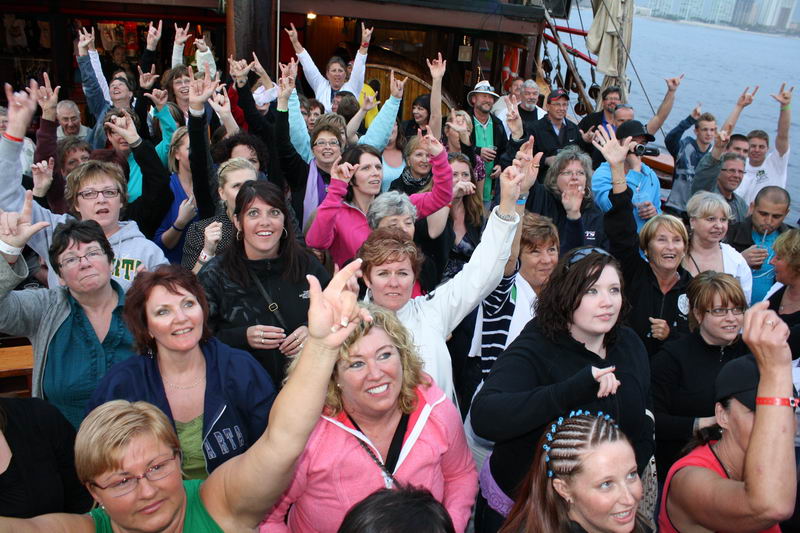 Large group of cheering passengers, mostly adults, crowded on the deck of the pirate ship at dusk, raising hands and rock-on gestures toward the camera. Rigging and city skyline faint in the background. Lively party atmosphere.
