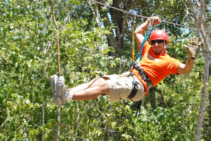 Helmeted visitor in an orange shirt gliding on a canopy zipline through the green jungle at Aktun Chen park, 