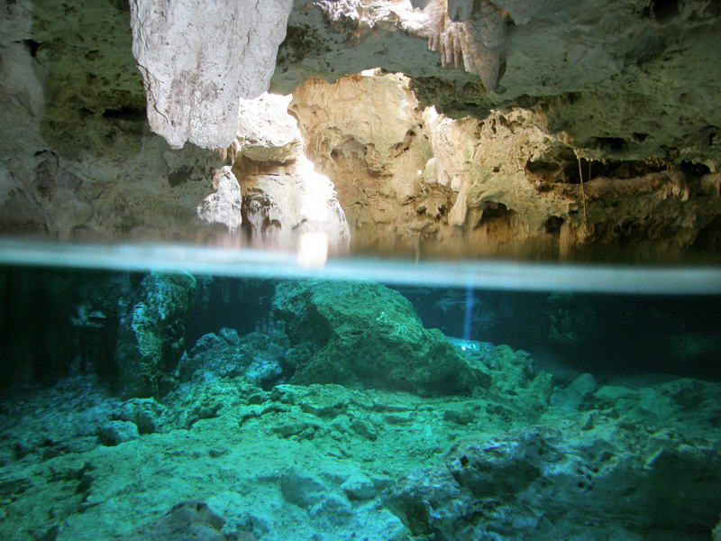 Half-submerged view of the Aktun Chen cenote showing rocky cave formations above and clear green water below the surface