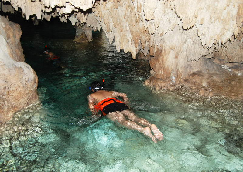 Snorkeler in an orange life vest swimming through a narrow stalactite passage in the Aktun Chen underground cenote