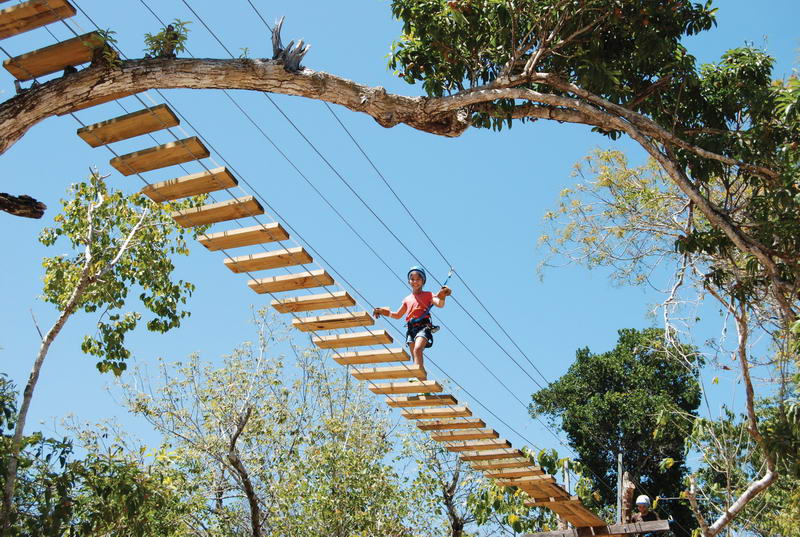 Child crossing a wooden suspension bridge strung between jungle trees on the Aktun Chen canopy course under a blue sky