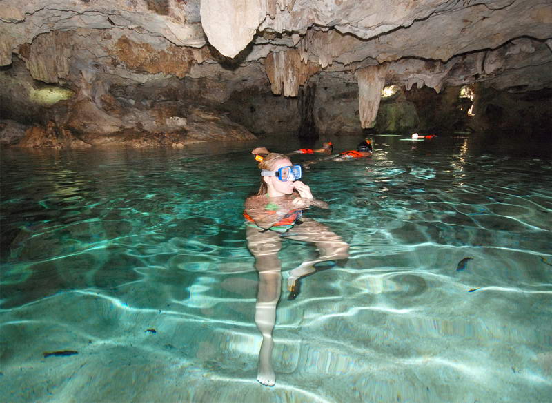 Snorkeler floating in the crystal-clear turquoise water of the underground cenote at Aktun Chen cave, stalactites overhead