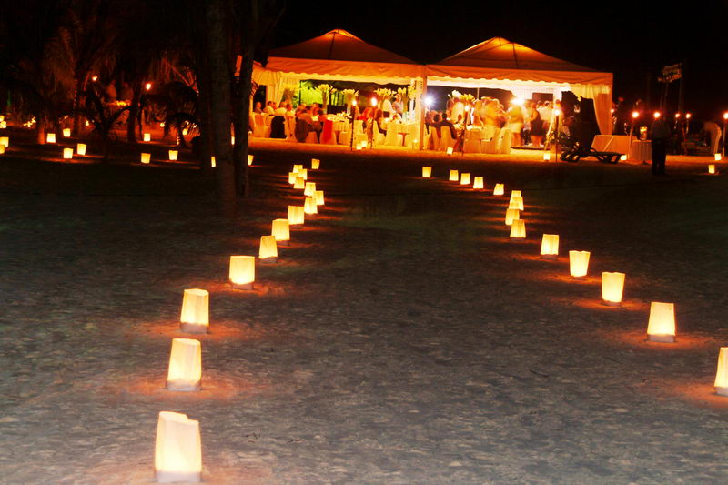 Nighttime beach reception with paper-bag candle luminaries lining a sandy path that leads toward two brightly lit white catering tents filled with guests.