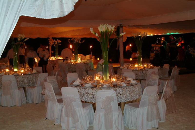 Elegant evening reception under a white tent: round tables with white ruffled chair covers, lace overlay cloths and tall calla-lily centerpieces lit by candlelight, with a band setting up at right.