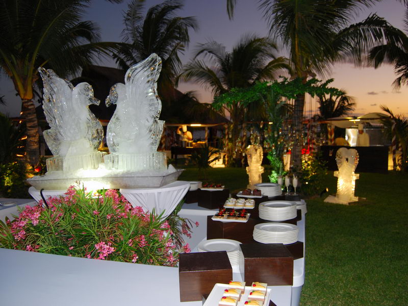 Outdoor dusk buffet featuring two facing swan ice sculptures on a white station, rows of stacked plates and dessert bites, with pink flowers, palm trees and an orange sunset sky behind.