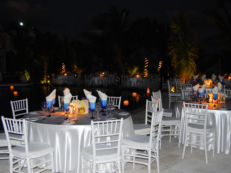 Nighttime beachside reception beside a pool or lagoon with round tables in white satin cloths, blue glassware and white chiavari chairs. Warm lit palm trunks and a moon appear in the dark background.