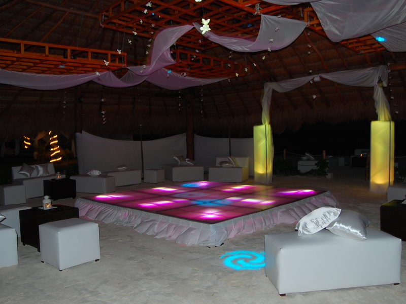 Beach lounge set up under a thatched palapa at night with an illuminated pink-and-purple LED dance floor at the center, surrounded by white lounge sofas and ottomans, draped white fabric and column lights.