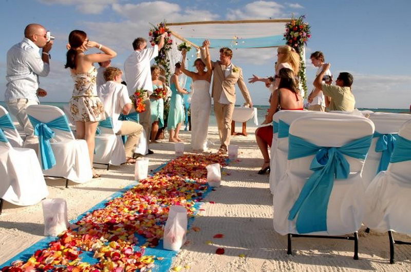 Joyful beach wedding recessional: a bride and groom walk down the aisle with arms raised in celebration while guests cheer, chairs tied with turquoise sashes and a bright petal-strewn aisle runner leading from a floral arch.