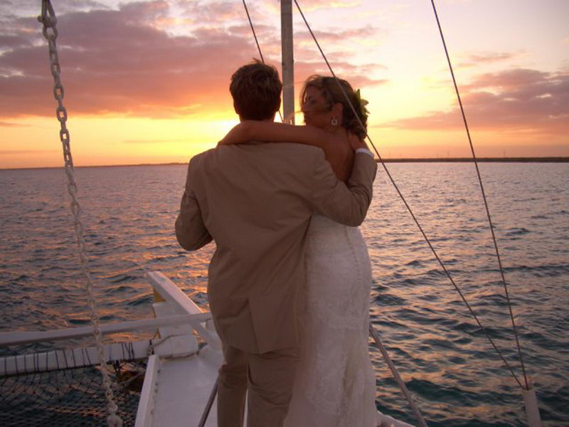 Rear view of a bride in a lace gown and groom in a tan suit embracing on the deck of a sailboat at sunset, looking out over calm water under a pink-and-orange sky.