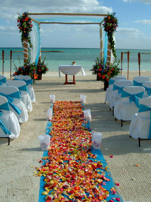 Beach wedding ceremony setup with a wooden arch topped by vivid multicolored flowers and turquoise drape, white chairs tied with blue sashes and a colorful petal aisle runner, facing the turquoise sea on a sunny day.