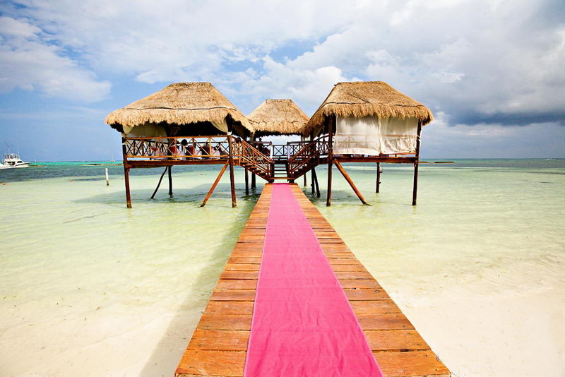 Overwater thatched palapa cabanas on wooden stilts, reached by a pier with a pink runner carpet leading across shallow turquoise water under a cloudy sky, a boat moored to the left.