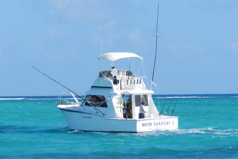 A white sportfishing boat named 'Moon Shadows II' with a flybridge and outriggers underway on turquoise water, anglers aboard, with pelicans perched on rocks to the right.