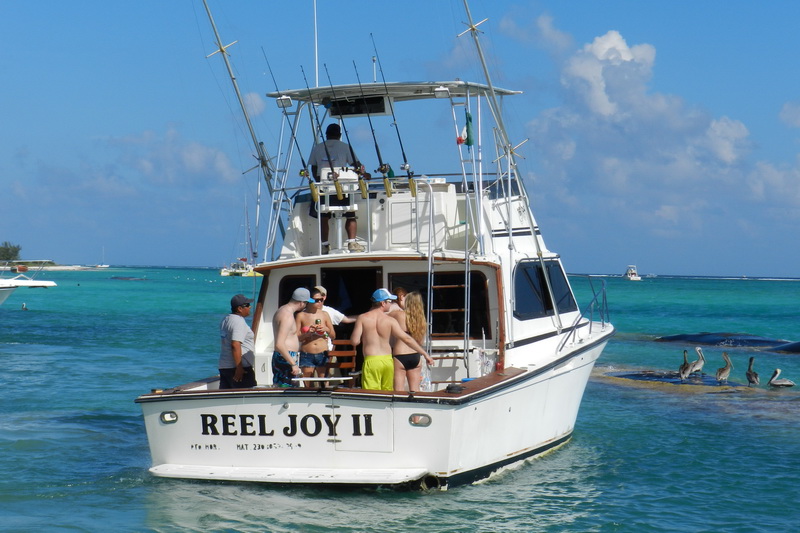 Stern view of a white sportfishing boat named 'Reel Joy II' with several young people aboard in swimwear, on turquoise water with pelicans resting on rocks to the right.