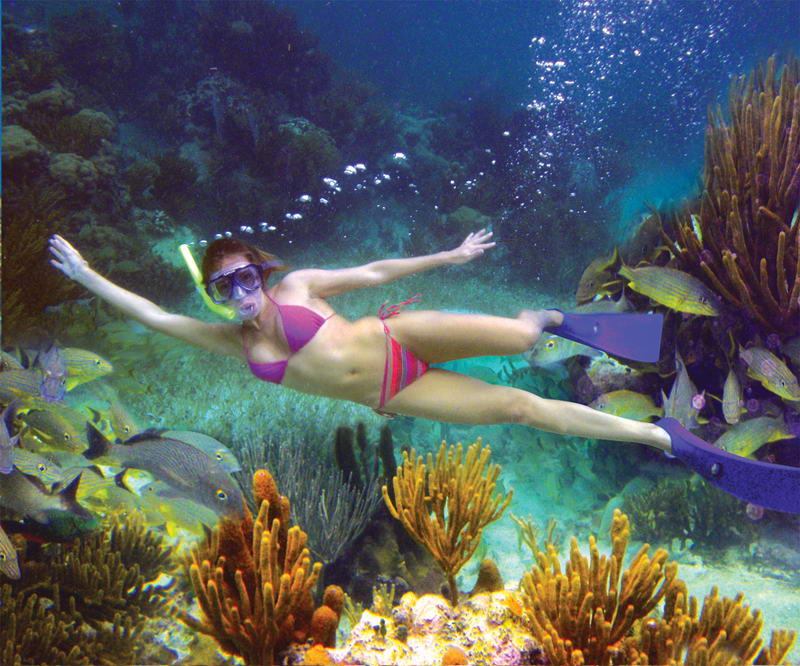 Underwater photo of a woman in a pink bikini and yellow snorkel mask gliding with arms outstretched over a vibrant coral reef, surrounded by schools of fish, orange sponges and gorgonians in clear turquoise water.