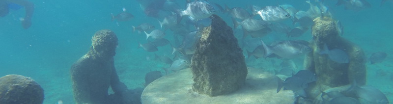 Escena de snorkel bajo el agua con las estatuas sumergidas del MUSA y un banco de peces plateados sobre el fondo arenoso frente a Cancún