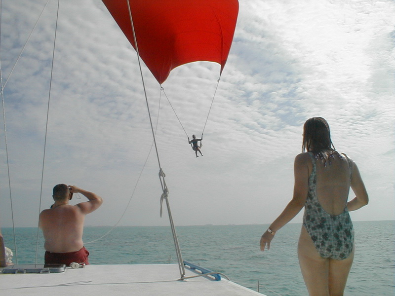 Aboard a sailing catamaran, a person swings on a line beneath a large red spinnaker sail while a shirtless man photographs and a woman in a patterned swimsuit watches from the deck. Overcast sky and calm sea behind.