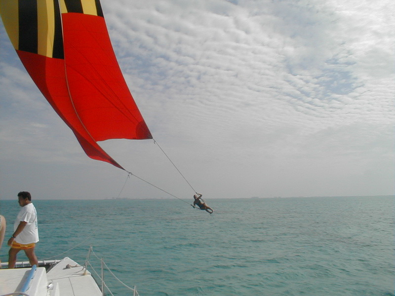 A person swings on a rope from a red, black and yellow striped spinnaker sail off a catamaran, feet skimming the sea, while a man in a white shirt watches from the deck. Cloudy sky over turquoise water.