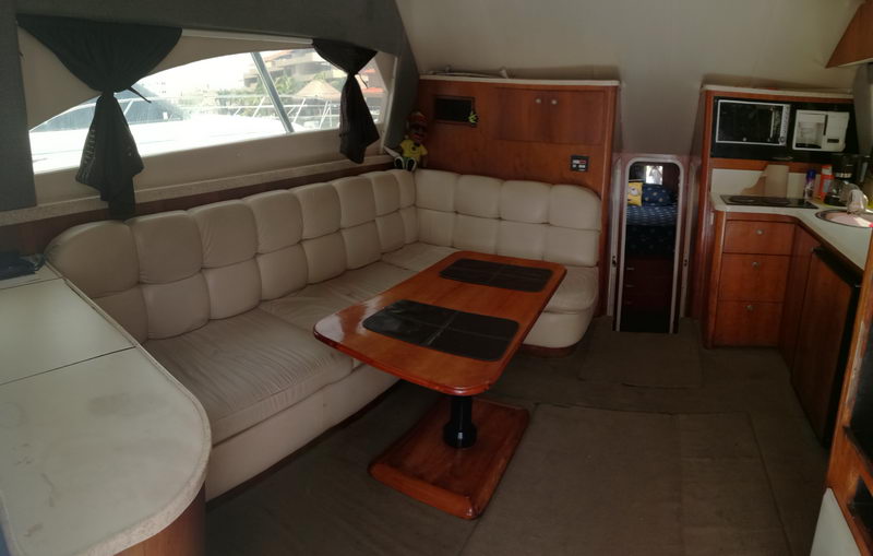 Interior salon of a sport-fishing boat with a cream leather L-shaped settee, a wood dining table with dark inlays, wood cabinetry and a compact galley to the right. A curtained window and passage to a forward cabin are visible.