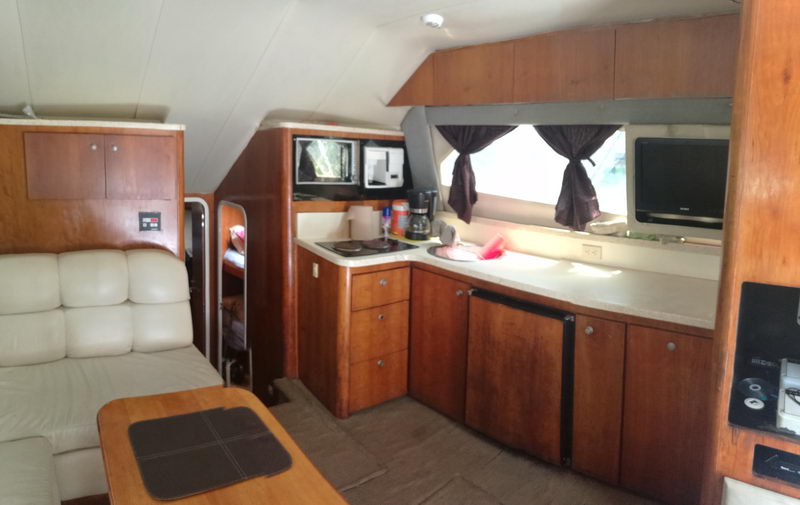 Interior galley and salon of a sport-fishing boat with wood cabinetry, a white countertop with a cooktop and coffee maker, a small TV, curtained windows and a cream leather settee with a wood table in the foreground.