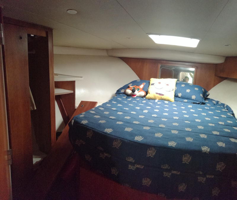 Interior cabin of a boat with a large double berth dressed in a blue crown-patterned bedspread, two pillows and a plush toy, flanked by wood-trimmed hull sides, a hanging locker and an overhead hatch light.