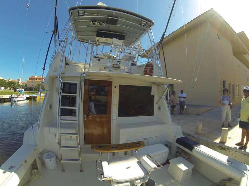 GoPro wide-angle stern view of a white sportfishing convertible at a dock, cockpit with fighting chair and coolers, wood cabin door and boarding ladder. Several people stand on the concrete dock beside the building under bright sun.