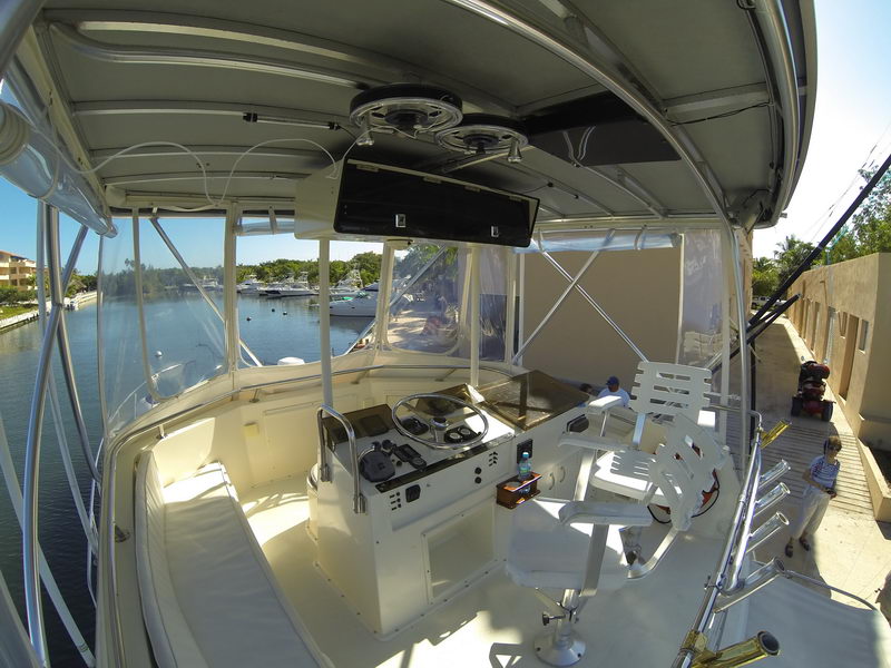 Wide-angle view of the open flybridge helm of a sportfishing yacht, showing the center console with wheel and electronics, a white helm chair and clear enclosure. Marina, docked boats and people on the dock are visible beyond under bright sun.