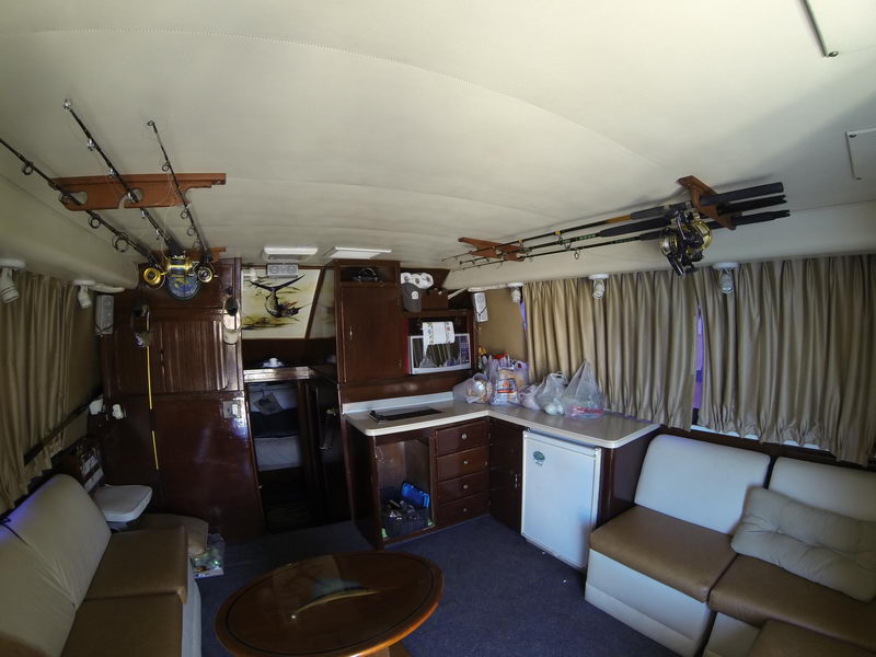 Interior salon of an older sportfishing yacht with beige and tan leather sofas, a round wood coffee table, wood cabinetry and a galley area with a mini-fridge. Fishing rods and reels are stored on overhead racks; a forward berth is visible.