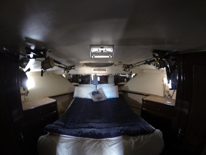 Dimly lit forward V-berth cabin of a sportfishing yacht with a navy-covered double bed flanked by nightstands, fishing rods and reels mounted on the overhead rails. Portholes and a small overhead light illuminate the cramped cabin.