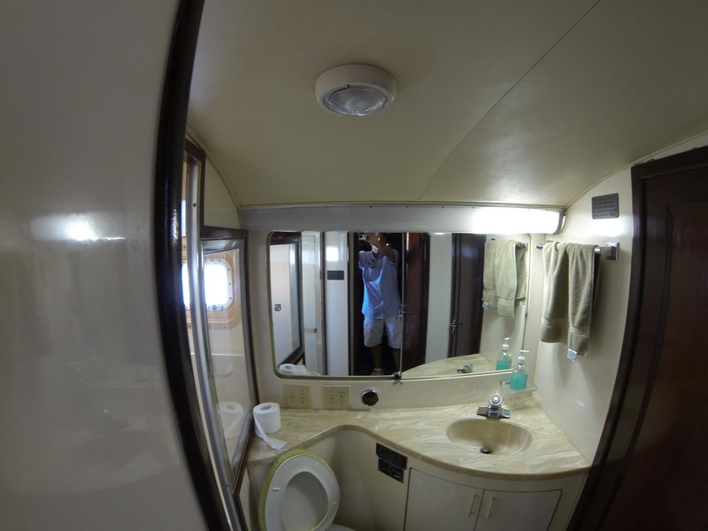 Cream-colored yacht head (bathroom) with a marble-look vanity, molded sink, mirror, marine toilet and folded towels. The photographer is reflected in the mirror. Wide-angle GoPro shot of the compact onboard bathroom.