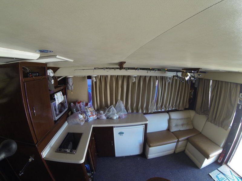 Salon and galley of a sportfishing yacht with a tan leather settee, wood cabinetry, a microwave, mini-fridge, cooktop and a curtained window band. Fishing rods hang overhead; grocery bags rest on the counter in the lived-in interior.
