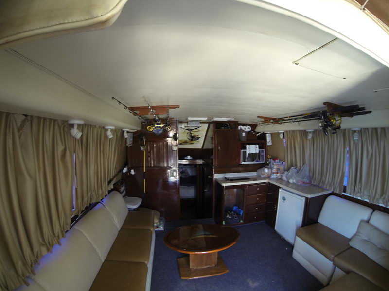 Wide-angle interior of a sportfishing yacht salon with beige leather sofas on both sides, an oval wood coffee table, curtained windows and a compact galley. Fishing rods and reels line the overhead racks in this lived-in cabin.