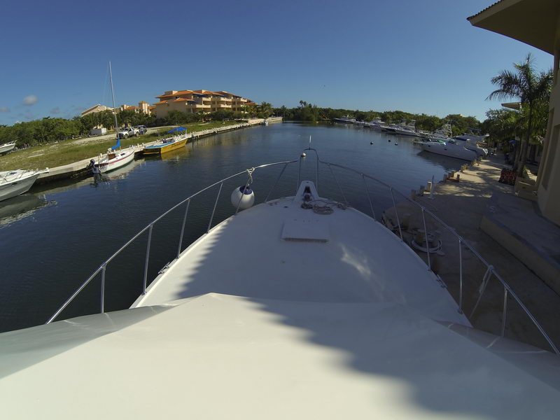Bow-forward GoPro view from a sportfishing yacht looking down a calm marina channel, the white foredeck and bow rail in the foreground. Docked boats, small pangas, waterfront condos and palms line the sunny channel under a clear blue sky.