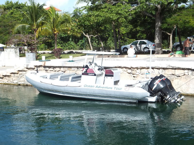 Grey rigid inflatable boat named Ocean Adventure Tour moored along a stone canal wall in a marina, with twin outboard engines, a center console T-top, palm trees and parked cars on the quayside behind.