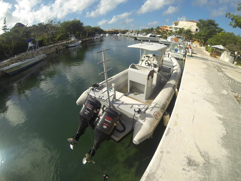 Fisheye view of a grey RIB with twin outboard engines and a center-console T-top moored at a marina dock, other sportfishing boats and residential buildings in the background under a blue sky.