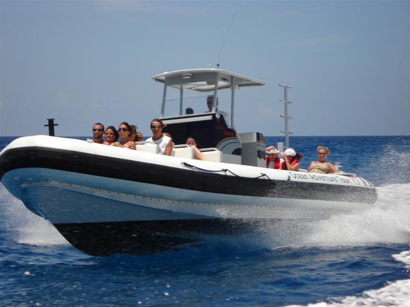 White-and-black RIB speedboat with a center-console T-top and about eight passengers speeding across deep blue water, kicking up spray, some riders wearing sunglasses and life vests. Marked Ocean Adventure Tour.