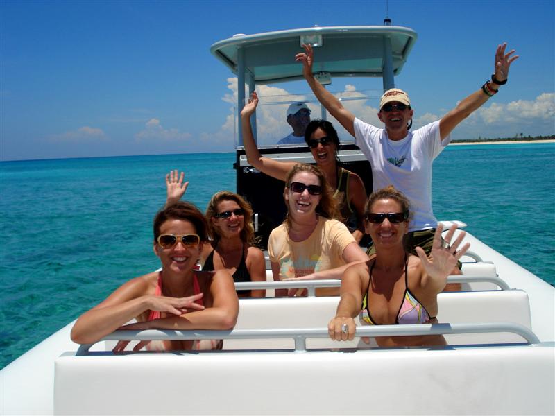 Group of six smiling women and men waving from the bow seating of a RIB speedboat, brilliant turquoise Caribbean water and a distant palm-lined shore behind them under a bright blue sky.