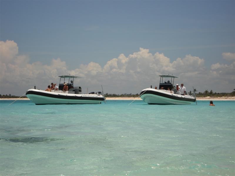 Two RIB speedboats anchored in shallow crystal-clear turquoise water off a white sandy beach, a swimmer floating nearby, palm-lined shore in the distance under a sky with scattered clouds.