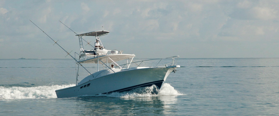 A white sportfishing boat with a dark hull cruising on open water under a cloudy sky, tuna tower with a person aboard and outriggers raised, seen from the bow quarter throwing spray.