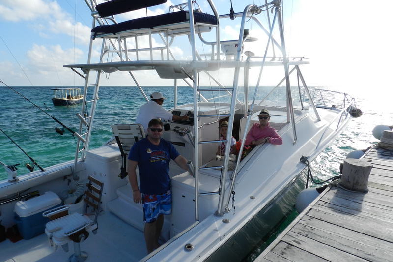 A white express sportfishing boat tied at a wooden dock with three anglers aboard in the cockpit and at the helm, tuna tower and outriggers overhead, calm turquoise sea and a small boat behind.