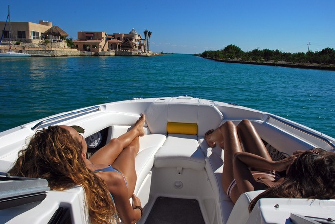 View from the bow of a boat cruising a calm channel: two tanned women in bikinis sunbathing on the white bow cushions, waterfront stone villas and green shoreline ahead under a deep blue sky.