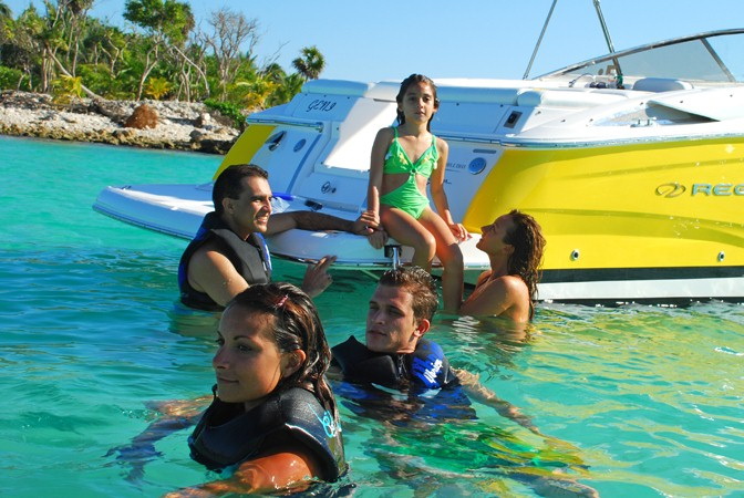 Family snorkeling in shallow turquoise water beside an anchored yellow Regal bowrider: three adults in the water wearing life vests and a young girl in a green swimsuit sitting on the boarding ladder, rocky tree-lined shore behind.