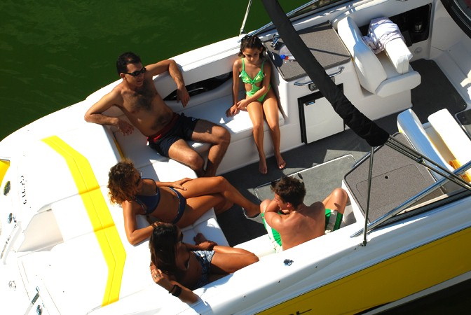 Elevated view into the cockpit of a yellow bowrider with six young people relaxing on white upholstered seats — some in swimwear talking and sunbathing — wakeboard tower shadow across the deck, dark green water alongside.