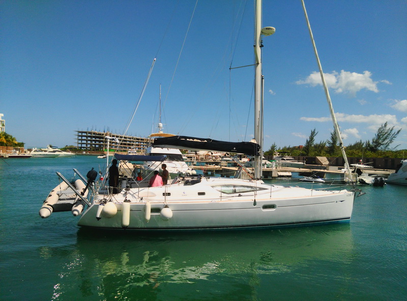 White Jeanneau sailboat with furled sails docked in a marina channel, gray inflatable dinghy hoisted on the port side, several guests boarding, other motor yachts, a catamaran and a building under construction behind.