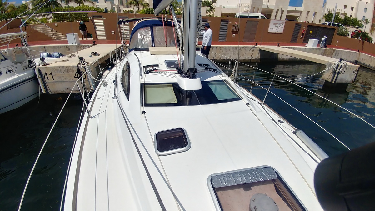 Looking forward along the white deck of a sailboat maneuvering beside numbered concrete marina docks, deck hatches and mast base in view, crew member in white shirt standing by the shrouds, fishing boat alongside.