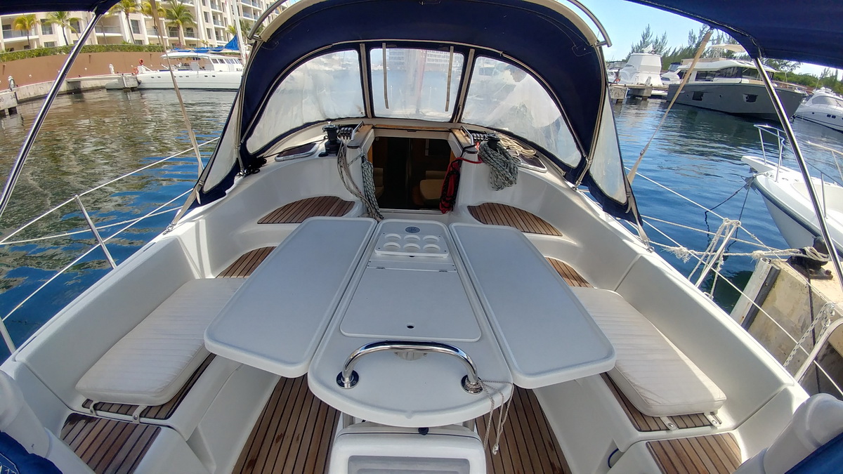 Cockpit of a sailboat seen from the helm: teak sole, cream cushioned benches on both sides, twin-leaf folding table with cup holders, navy bimini and clear-panel sprayhood, marina and boats around.