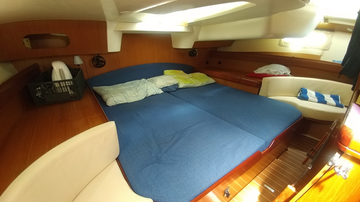 Aft owner's cabin of a sailboat with a wide double berth dressed in a blue quilted cover with pillows, warm wood joinery, cream upholstered bench seat with a striped towel and overhead hatches.