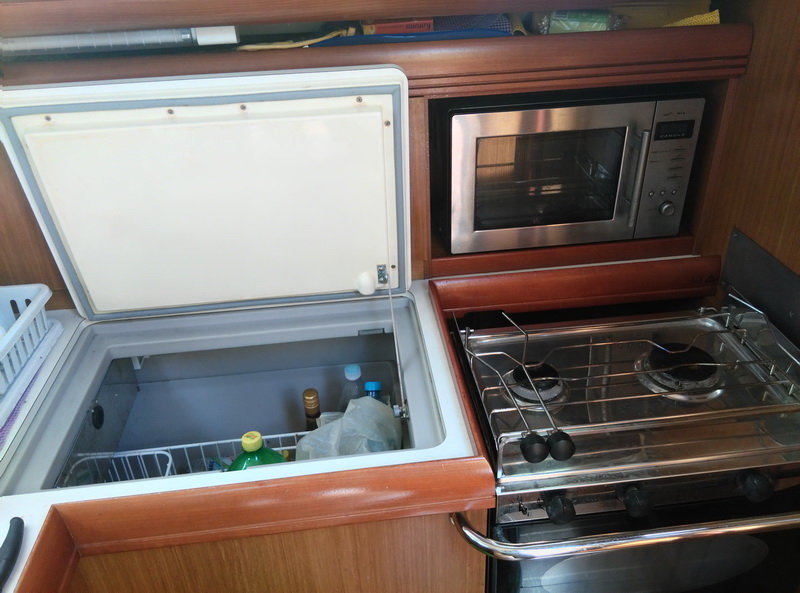 Sailboat galley interior with top-loading fridge open showing drinks, stainless microwave oven in wood cabinet, and two-burner gimbaled stainless stove, all set in warm wood joinery.